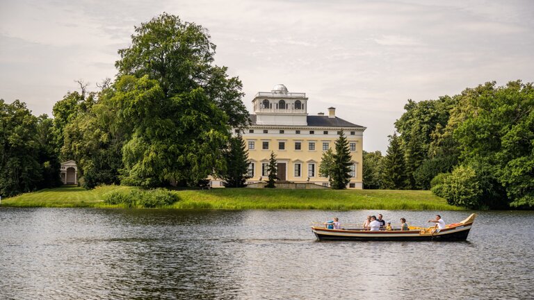 Foto von einem kleinen See, auf dem ein Boot mit Menschen fährt, im Hintergrund steht ein großes Haus umgeben von Bäumen | © Investitions- und Marketinggesellschaft Sachsen-Anhalt mbH / Nilz Böhme