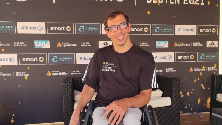 Johannes Grasser sitzt lächelnd in seinem Rollstuhl vor dem Werbebanner der deutschen Sporthilfe. | © Johannes Grasser