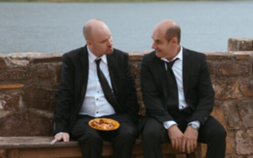 Aufnahme eines Filmausschnitts: links Igor mit einem Teller auf dem Schoß und rechts Lous, sitzen auf einer Bank an einem Fluss und blicken sich an. | © X Verleih