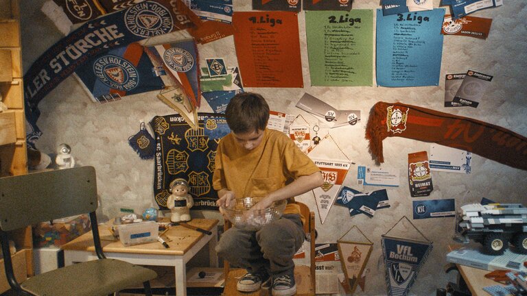 Jason in seinem Zimmer vor einer Wand mit Fanschals. Er hockt auf einem Stuhl mit einer Schale, darin Zettel mit allen Fußballvereinen in Deutschland. | © Szenenbild Wochenendrebellen I JETZT & MORGEN GbR
