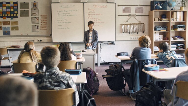 Jason (Cecilio Andresen) steht an der Tafel vor seinen Mitschüler*innen | © Szenenbild Wochenendrebellen I JETZT & MORGEN GbR