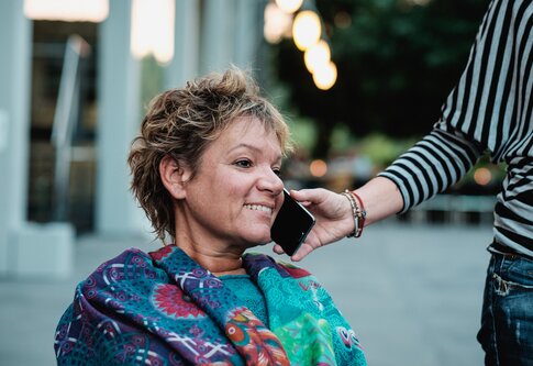 Assistenzperson hält einer Frau mit einer Behinderung das Handy ans Ohr. | © Andi Weiland/Gesellschaftsbilder.de