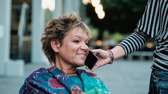 Assistenzperson hält einer Frau mit einer Behinderung das Handy ans Ohr. | © Andi Weiland/Gesellschaftsbilder.de