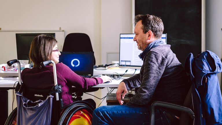 Zwei Personen sitzen am Schreibtisch, auf dem zwei Computer stehen. Eine Person sitzt im Rollstuhl und die Personen sind einander zugewandt. | © Andi Weiland/ Gesellschaftsbilder.de