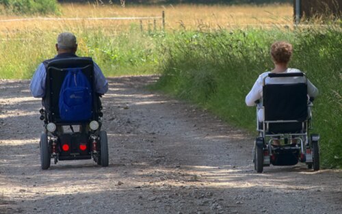  zwei Personen fahren nebeneinander auf einem Schotterweg mit einem elektrischen Rollstuhl. Das Bild ist von hinten aufgenommen, es ist ein schöner Tag. | © pixabay.com