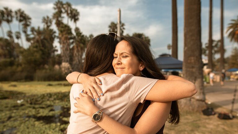 Auf dem Bild sieht man zwei Frauen, die sich fest umarmen. Im Hintergrund sieht man Palmen. | © Pexels.com