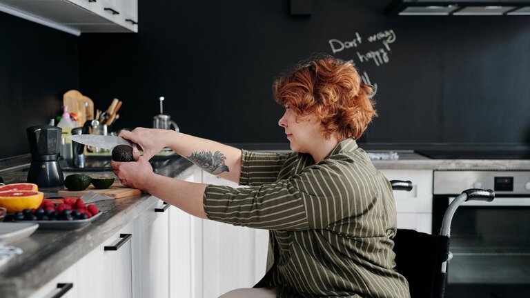 In einer modern eingerichteten Küche ist eine Frau mit roten Haaren im Rollstuhl. Sie schneidet mit einem Messer eine Avocado in zwei Hälften. Auf der Arbeitsplatte steht auch eine Espressomaschhine und diverses Obst. | © Marcus Aurelius / Pexels.com