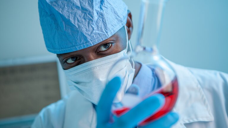 Foto von einem Mann, der Arztkleidung und eine medizinische Maske trägt, in seiner Hand hält er ein Reagenzglas, das mit einer roten Flüssigkeit gefüllt ist. | © Unsplash