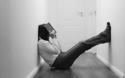 Eine junge Frau sitzt im Wohnungflur zwischen zwei Wänden und hält ein Buch über ihr Gesicht | © unsplash