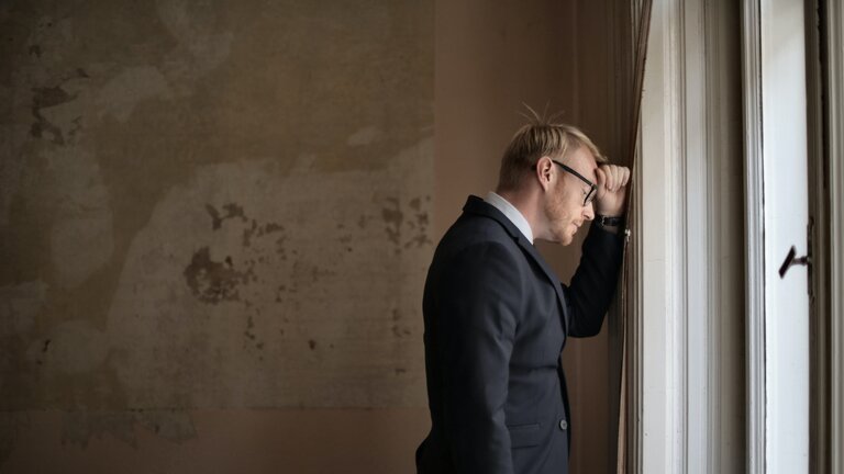 Ein Mann im Anzug lehnt verzweifelt an einer Wand am Fenster | © Andrea Piacquadio / pexels