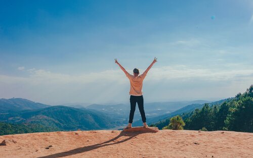 Eine Person steht auf der Spitze eines Berges mit ausgestreckten Armen – die Hände formen Peace-Zeichen | © Min An / pexels