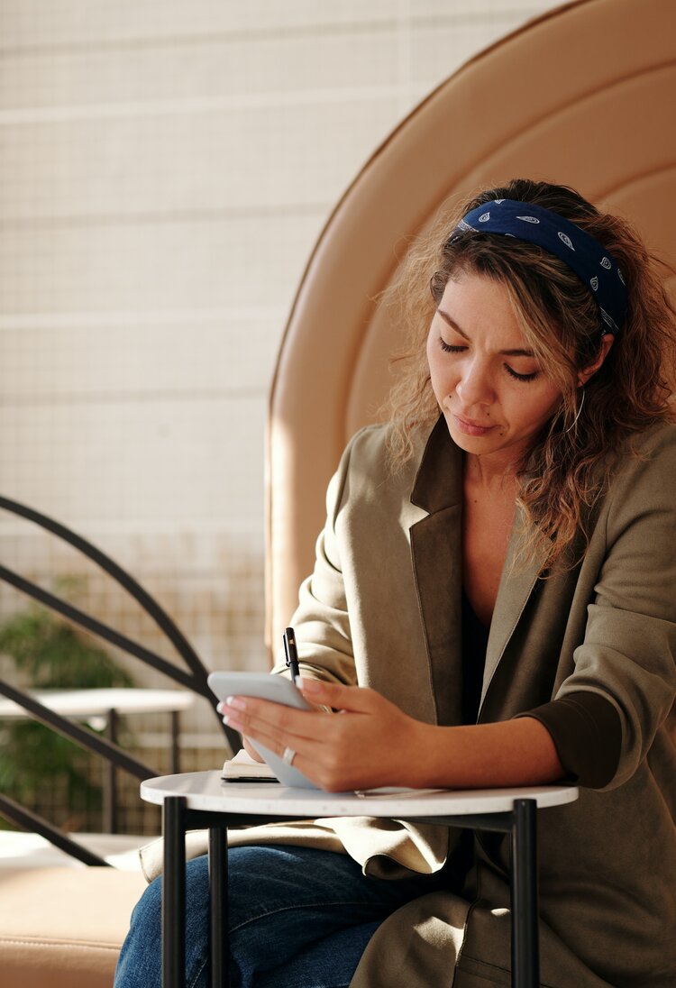 Eine Frau sitzt an einem Coffee Table, macht sich Notizen und hält dabei ein Smartphone in der Hand | © Marcus Aurelius / pexels