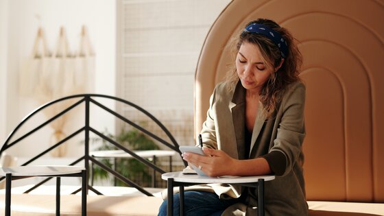 Eine Frau sitzt an einem Coffee Table, macht sich Notizen und hält dabei ein Smartphone in der Hand | © Marcus Aurelius / pexels