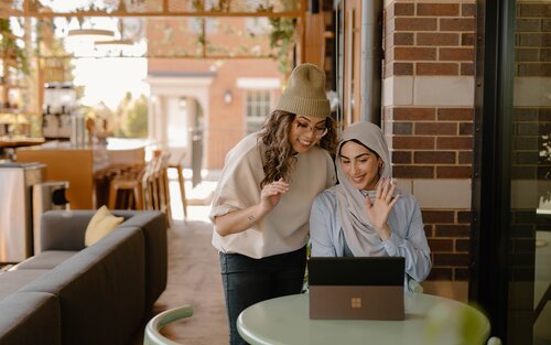 Zwei Frauen winken in die Webcam eines Laptops. Die eine steht hinten und trägt einen hellen Pulli, eine schwarze Hose und eine Mütze, die andere sitzt vorne und trägt eine hellblaue Bluse und ein helles Kopftuch. | © Geeignete Berufe für Menschen mit Depressionen