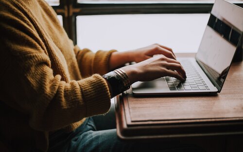 Oberkörper einer Person mit gelb-braunem Wollpullover an einem Tisch sitzend, die Hände auf einer Laptop-Tastatur. | © Christin Hume / Unsplash