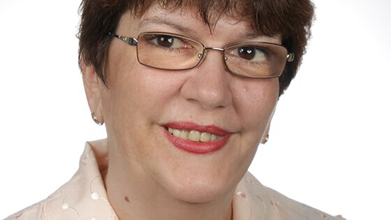 Portraitfoto einer Frau mit kurzen Haaren und Brille, die eine helle Bluse und eine Kette trägt.