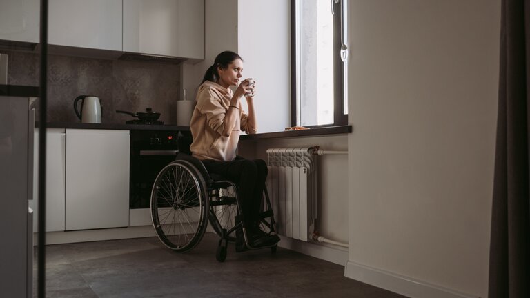 Eine Frau im Rollstuhl mit einer Tasse in der Hand sieht zum Küchenfenster hinaus  | © pexels