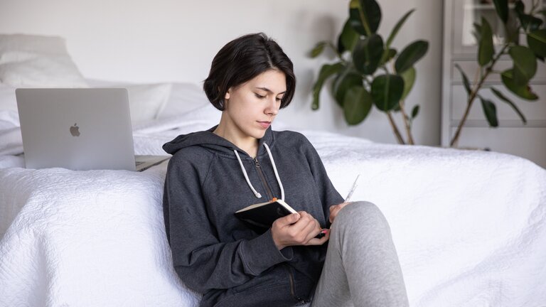 Eine Frau sitzt an ihrem Bett und schreibt in ein Notizbuch. | © pexels