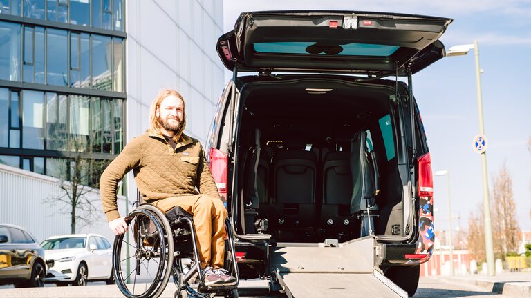 Ein Mann im Rollstuhl sitzt neben einem Auto. Das Auto hat eine offene Heckklappe und eine Rollstuhlrampe führt in das Auto. | © Andi Weiland/ Gesellschaftsbilder.de