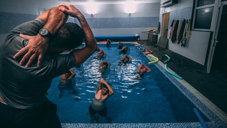 Eine Person dehnt steht am Beckenrand seinen Arm während mehrere ihm zugewandt im Schwimmbecken die gleiche Übung machen. | © unsplash
