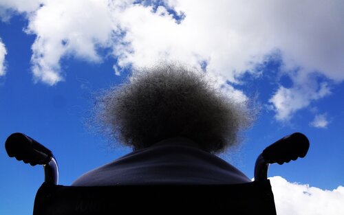 Man sieht Rücken von Mann in Rollstuhl und blauen Himmel mit ein paar Wolken. | © James Williams/ unsplash