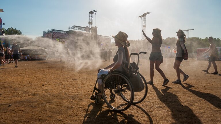 Eine Rollstuhlfahrerin bewegt sich auf einem staubigen Festivalgelände. Im Hintergrund sieht meine eine Bühne. | © Timo Hermann/ Gesellschaftsbilder.de