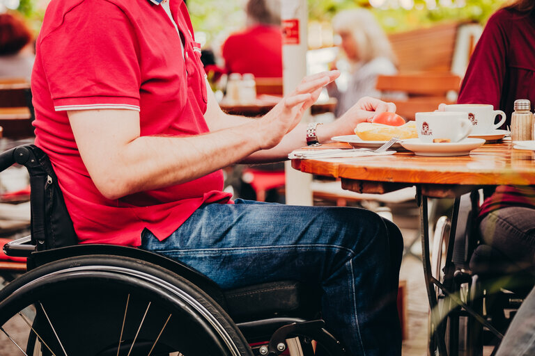 Eine Person im Rollstuhl sitzt draußen an einem Tisch in einem Café. | © Andi Weiland/ Gesellschaftsbilder.de