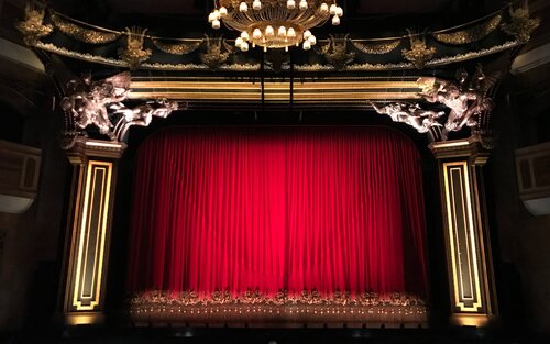 Im Mittelpunkt zu sehen, ist der rote Vorhang eines punkvollen Theaters, welches  an den oberen Seiten neben dem Vorhang Statuen hat. Vor dem Vorhang befindet sich ein großer Kronleuchter. | © Gwen King/ unsplash