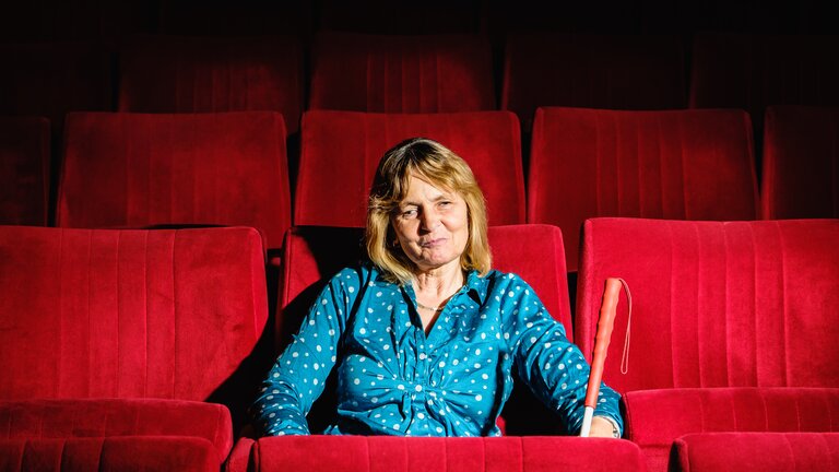 Frau sitzt auf einer roten Sitzbank im Kino oder Theater mit einem Blindenstock neben ihr. | © Andi Weiland /Gesellschaftsbilder.de