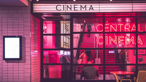 Blick von außen auf den Eingang eines Kinos, welches ein rotes Neonschild und rote Stufen hat. | © Myke Simon/unsplash