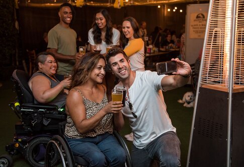 Eine Gruppe von Menschen, die gemeinsam in einer Bar sind und Spaß haben. Zwei Frauen sitzen dabei auch im Rollstuhl. | © unsplash