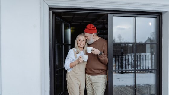 Ein Paar mittleren Alters steht in der Türschwelle einer Balkontür mit Kaffetassen in der Hand. | © pexels