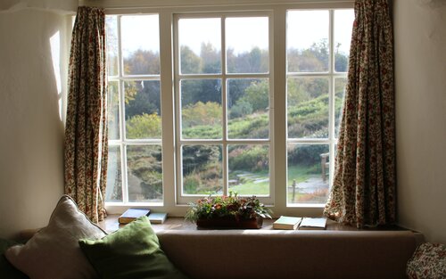 Ein weißes Fenster mit bunten Gardinen. Auf dem Fensterbrett befinden sich eine Pflanze und Bücher. Vor dem Fenster steht ein Sofa mit Kissen.  Durch das Fenster sieht man eine grüne Landschaft. | © Rob Wingate/unsplash