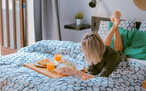 Eine Frau liegt mit dem Bauch auf dem Bett und hat neben sich ein Tablett mit Frühstück liegen. | © Toa Heftiba/unsplash