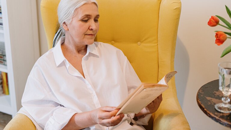 Seniorin sitzt in einem gelben Sessel und liest. | © pexels