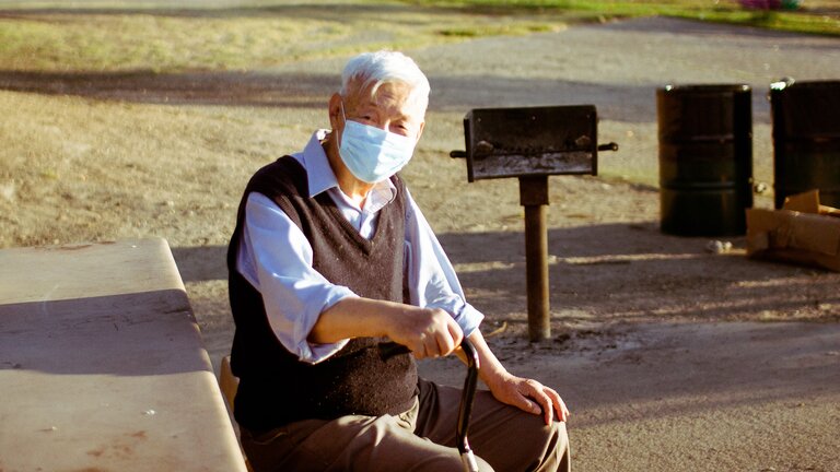 Ein älterer Herr mit Maske sitzt auf einer Parkbank. | © unsplash