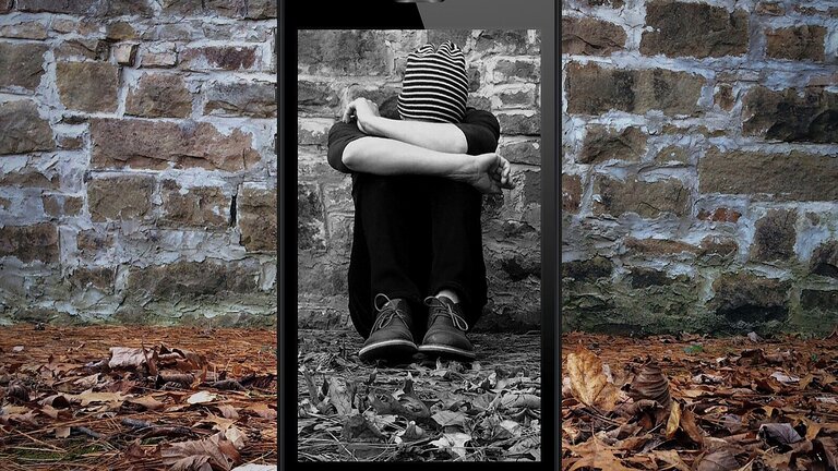 Foto von einem Menschen, der auf dem Boden sitzt und den Kopf auf den Armen aufstützt. Das Foto wird auf einem Smartphone in Schwarzweiß dargestellt. | © Pixabay