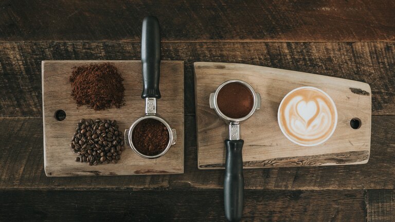 Kaffee wird auf einer Holzfläche in verschiedenen Formen präsentiert – als Pulver, als ganze Bohnen, im Siebträger und als Cappucino in einer Tasse. | © Nathan Dumlao/unsplash