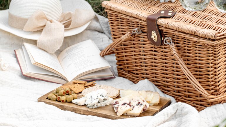 Auf einer Grasfläche liegt eine weiße Decke. Auf ihr befinden sich Picknickkorb, ein Sonnenhut, ein aufgeklapptes Buch und ein Holzbrett mit Käsescheiben, Oliven und Krackern. | © Evangelina Silina/unsplash
