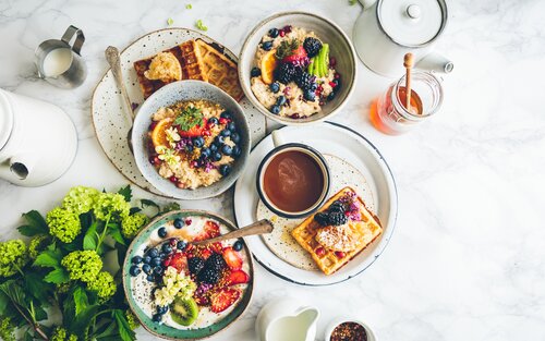Eine Aufnahme eines Tisches mit verschiedenen Frühstücksgerichten – Haferbrei mit Obst, Waffeln, Honig, Milchkännchen, Karaffen, Kaffee | © Brooke Lark/unsplash