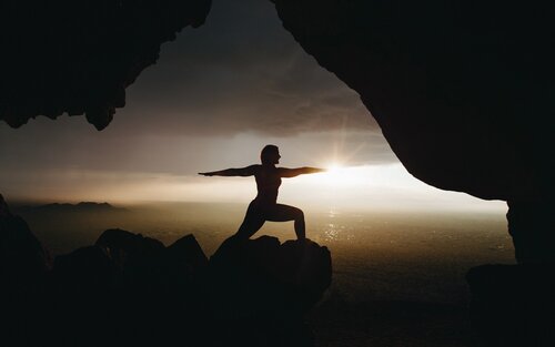 Draussen auf einem Stein vor dem Meer führt eine Person die Kriegerposition aus.  | © unsplash
