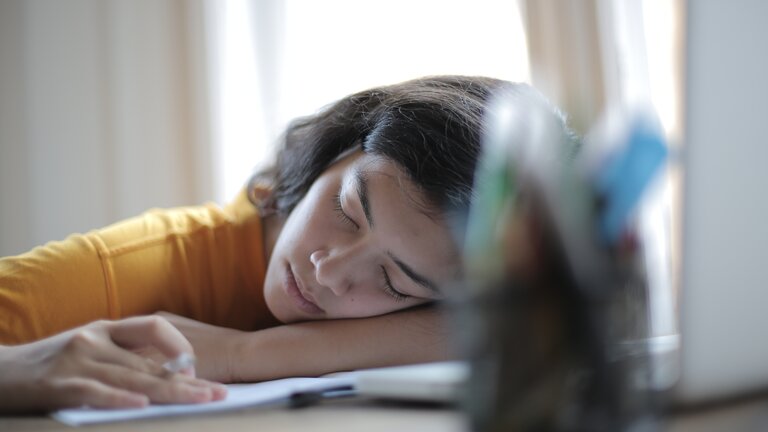 Eine junge Frau sitzt an ihrem Arbeitstisch, auf dem ein Notizbuch, ihr Laptop und ein Behälter mit Stiften stehen. Die Frau hat ihren Kopf auf ihren Arm und den Tisch gelegt und schläft.