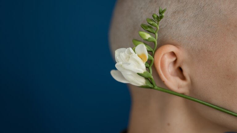 Nahaufnahme eines Kopfes von der Seite. Um das Ohr liegt eine weiße Pflanze mit grünem Stiel.