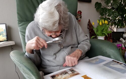 Eine ältere Dame sitzt auf einem Sessel und betrachtet mithilfe einer Lupe ein Fotoalbum. | © pixabay