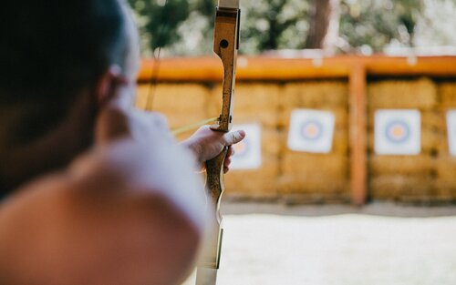 Im Fokus ist eine Person, die einen Bogen spannt. Im Hintergrund sind unscharf Zielscheiben zu sehen. | © Vince Fleming/unsplash