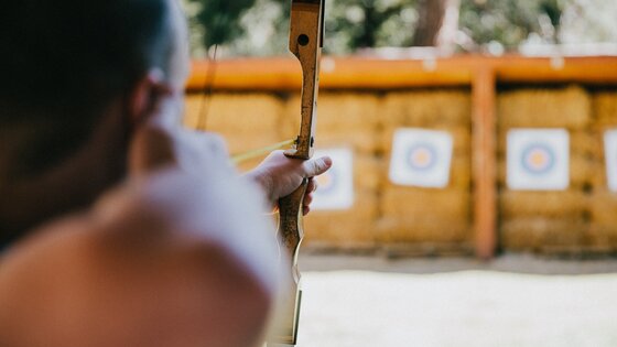 Im Fokus ist eine Person, die einen Bogen spannt. Im Hintergrund sind unscharf Zielscheiben zu sehen. | © Vince Fleming/unsplash
