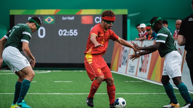 Drei Fußballspieler mit Augenbinden spielen Fußball. Im Hintergrund sieht man den Punktestand und die brasilianische und chinesische Flagge. | © Andi Weiland/ Gesellschaftsbilder.de