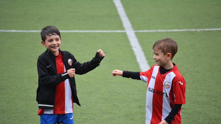 Zwei lachende Kinder mit rot-weißen Trikots stehen auf einem Fußballplatz. | © Adrià Crehuet Cano/ unsplash