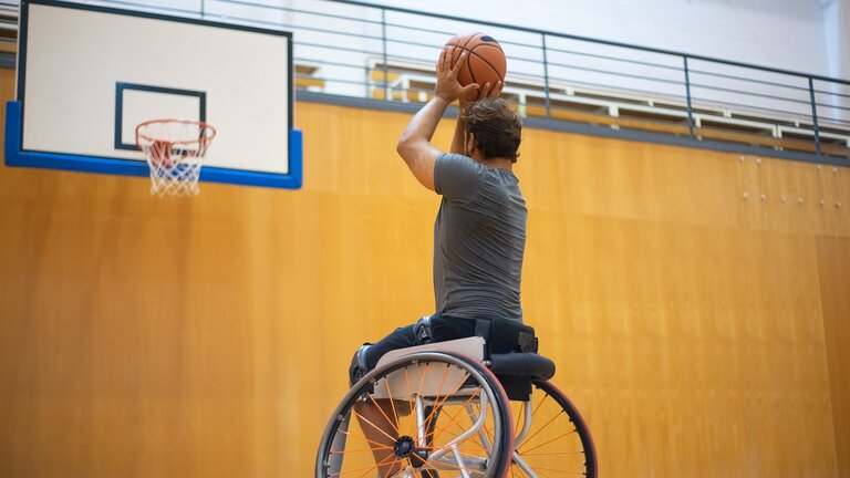 Ein Mann im Rollstuhl befindet sich in einer Sporthalle. In der Hand hält er einen Basketball und zielt mit diesem auf den Korb. | © Pexels.com