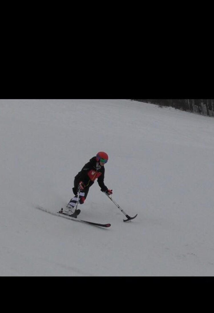 Eine Person mit amputiertem linkem Bein fährt auf Skiern eine Piste herunter, die Person trägt einen schwarzen Skianzug und einen roten Helm | © privat